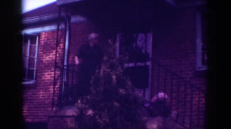 1962 Raleigh home-movie frame showing a man on a brick house porch at night, with shrubs, window trim, and metal railings visible.