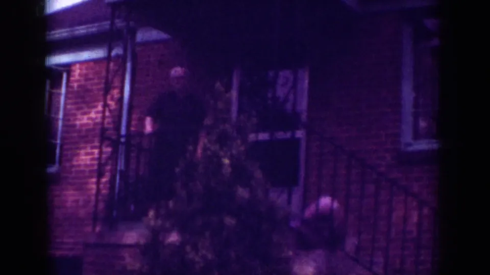 1962 Raleigh frame of the man still standing on the shadowed porch of a brick house, with railings and shrubs in the foreground.