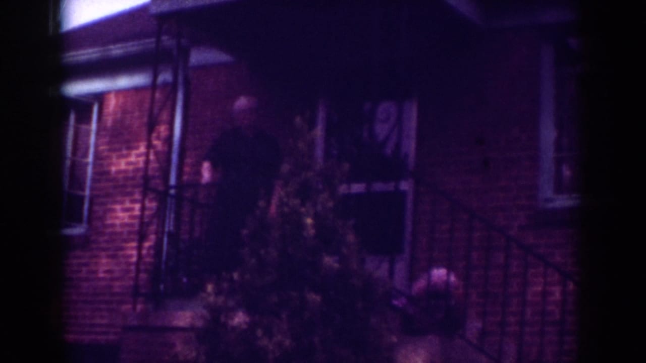 1962 Raleigh home-movie frame showing an older man on a brick house porch near a small evergreen tree and railing.