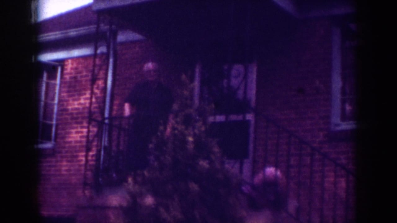 Older man on porch partially framed by a small tree and dark doorway.