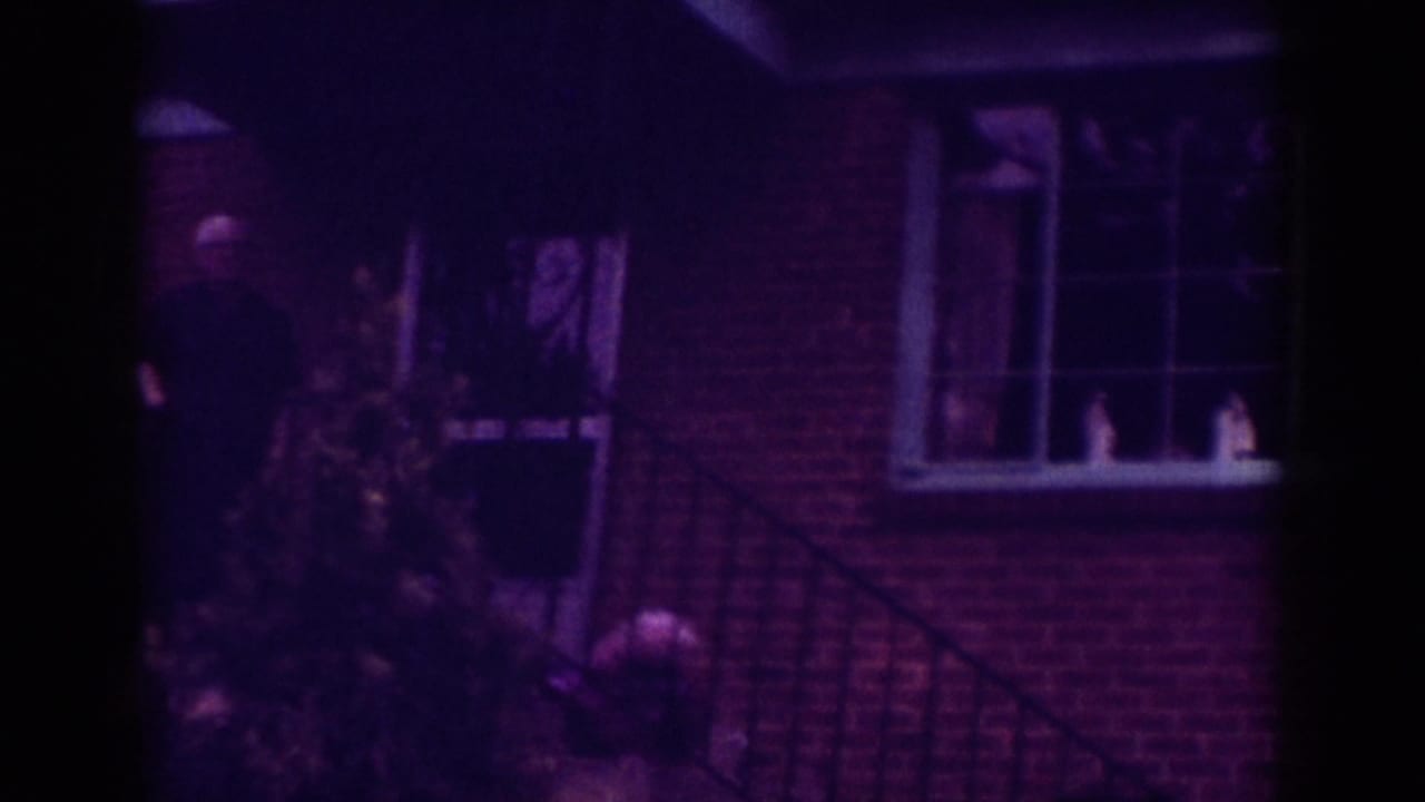 1962 Raleigh home-movie frame showing a brick house exterior with a window, porch railing, and shrub in dim purple-tinted light.