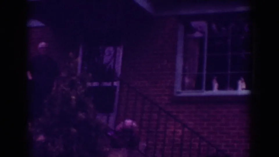 1962 Raleigh home-movie frame showing a brick house exterior with a window, porch railing, and shrub in dim purple-tinted light.