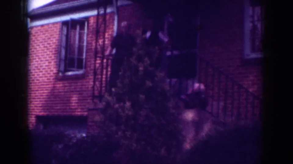 1962 Raleigh home-movie frame showing a dark-clothed person near porch steps, partly obscured by a shrub and railing.