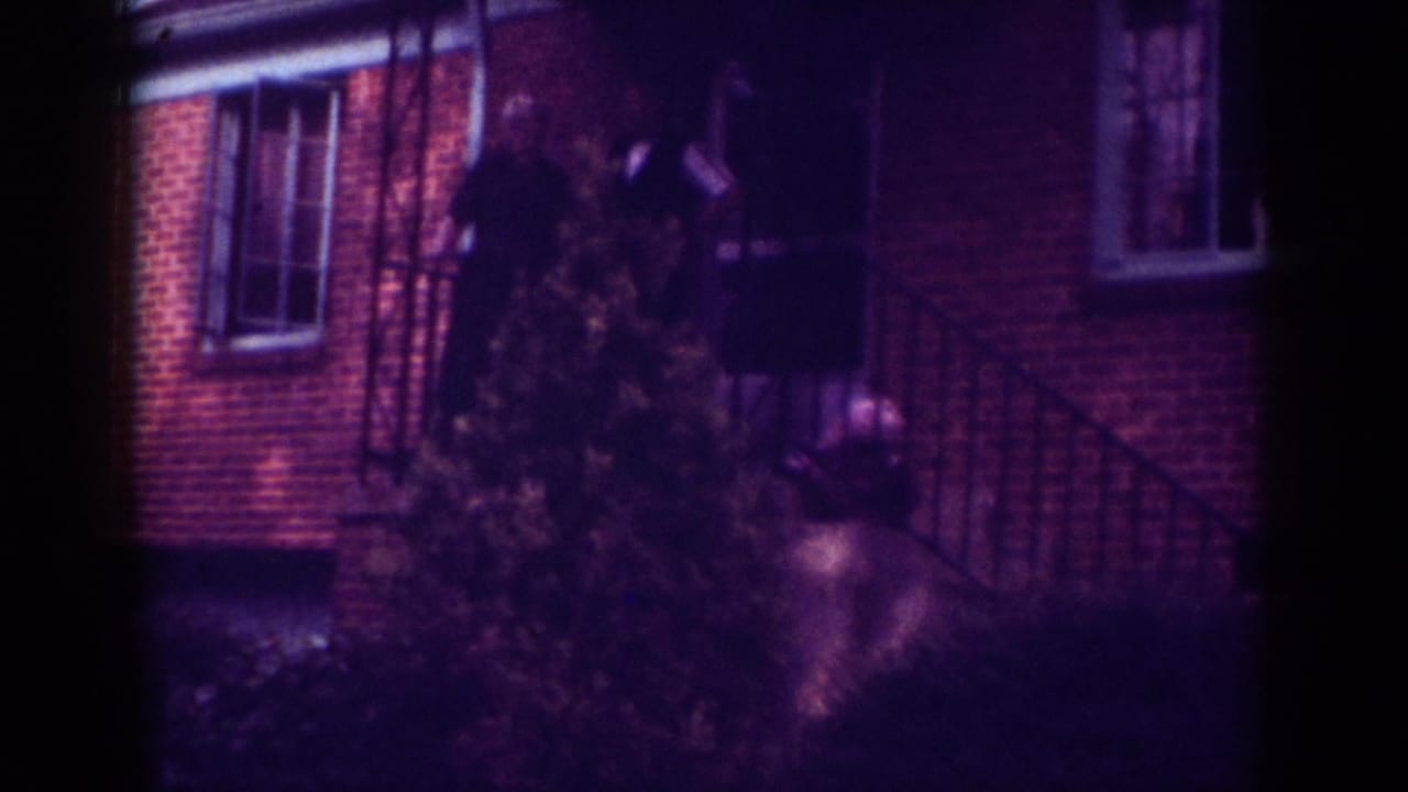 Raleigh, North Carolina 1962 home-movie frame showing two adults near a brick house entrance, partly obscured by foliage.