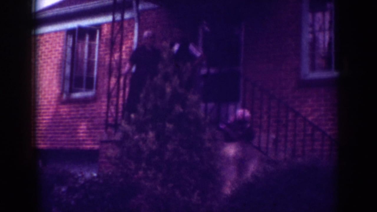 Adults stand near a brick house entrance behind a shrub and stair railing.