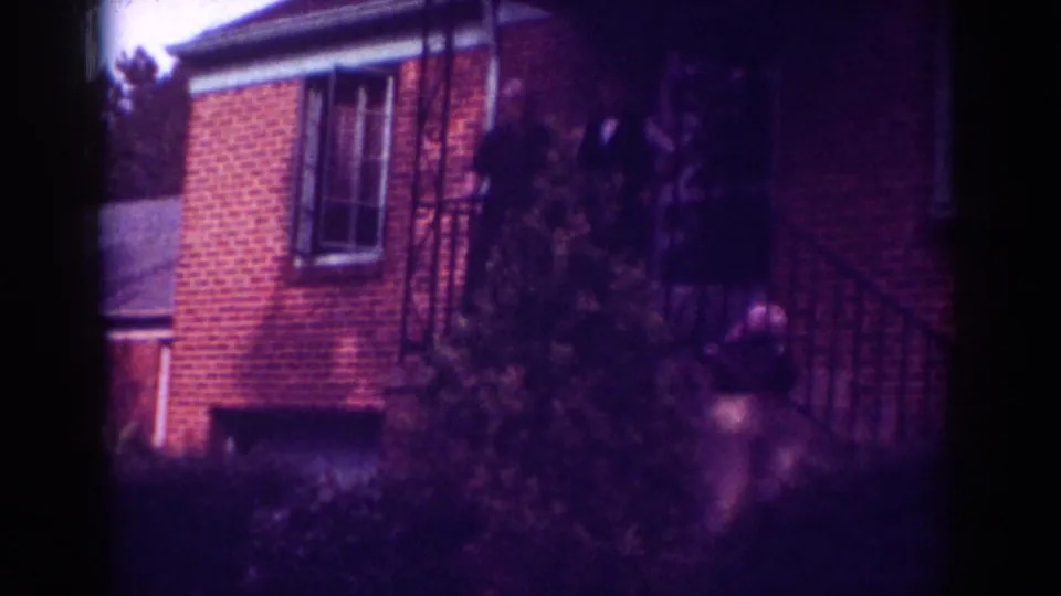 Raleigh, North Carolina 1962 frame showing adults at a brick house entrance, partly concealed by shrubbery in aged 8mm color.