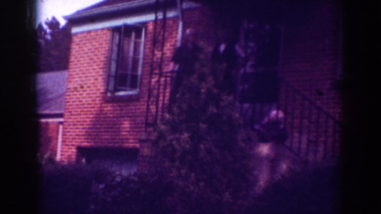 1962 Raleigh home-movie frame showing a red brick house, porch railings, greenery, and figures near the entrance.