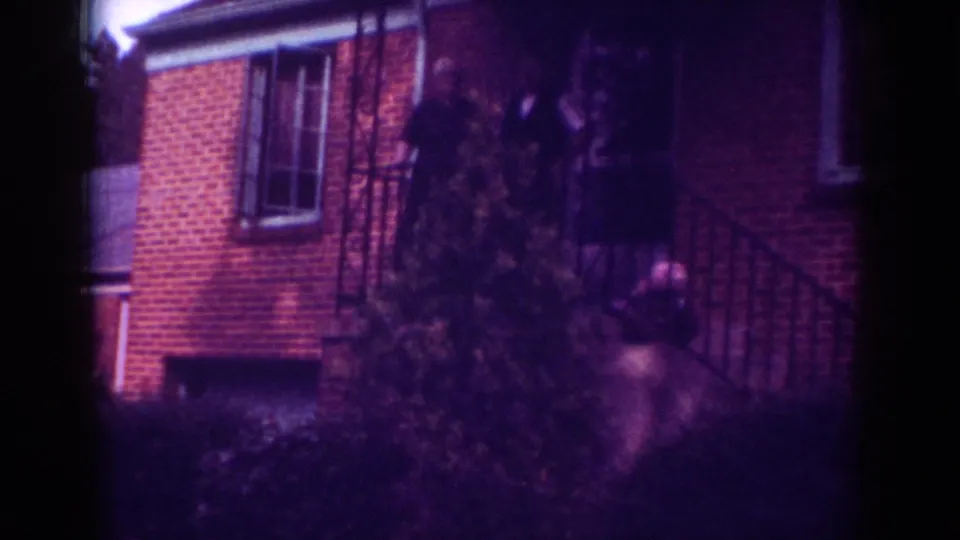 Aged 8mm frame from Raleigh in 1962 showing a brick house exterior and blurred people near the front porch.