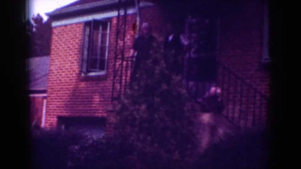 1962 Raleigh home-movie still showing people around the porch of a red brick residence with strong magenta film cast.