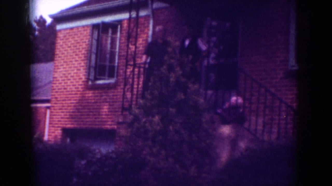 1962 Raleigh home-movie frame showing a brick house facade, porch area, shrubs, and indistinct people near the front steps.