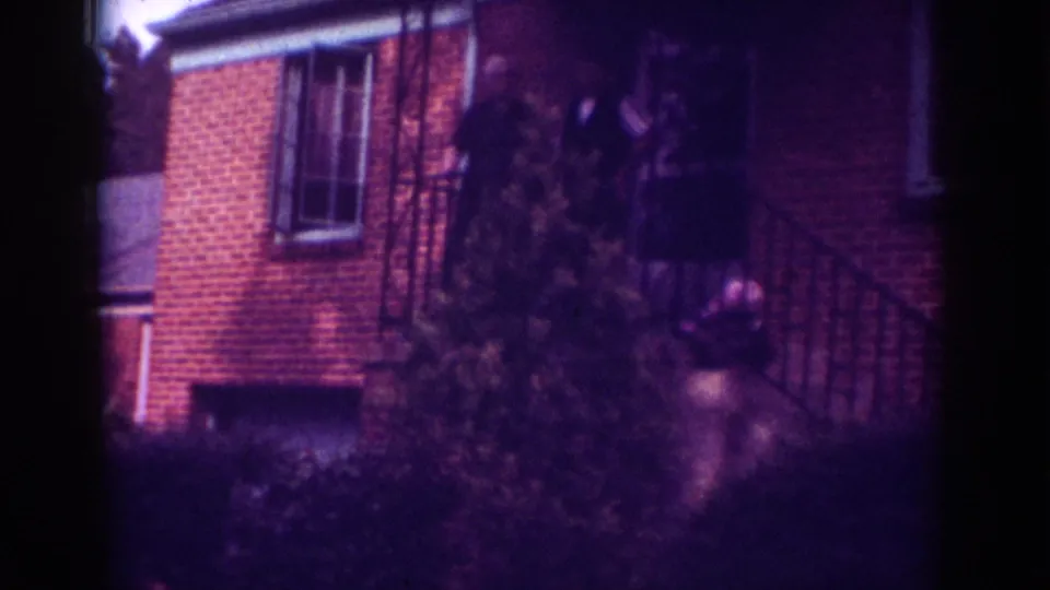 Home-movie view of a Raleigh brick house in 1962, with porch railings, landscaping, and indistinct figures near the entrance.