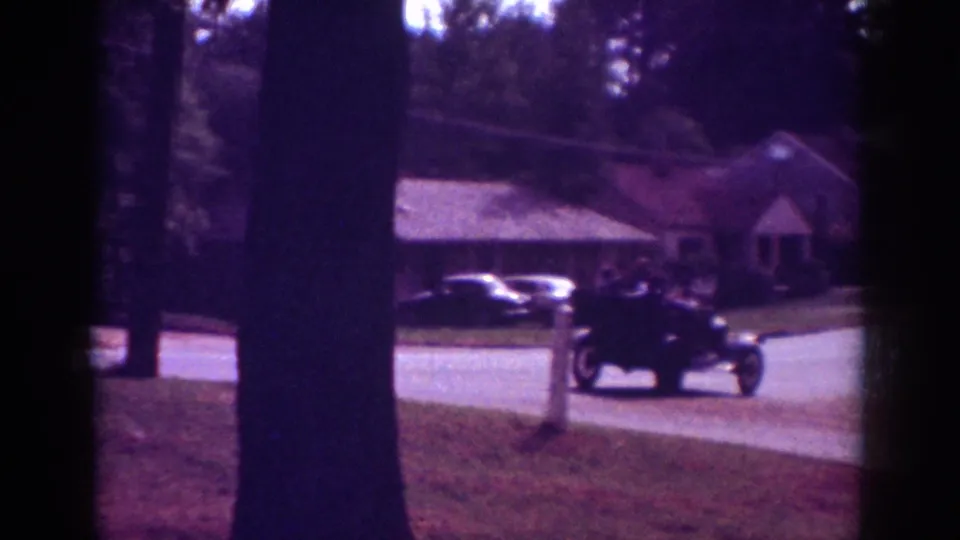 1962 Raleigh suburban street view with lawns, trees, nearby houses, and a dark classic car moving along the road.