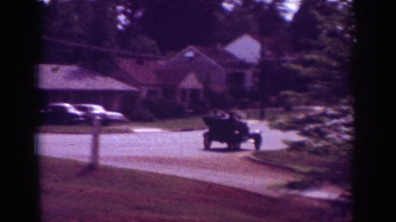1962 Raleigh home-movie frame showing a dark car on a tree-lined suburban street near brick houses.