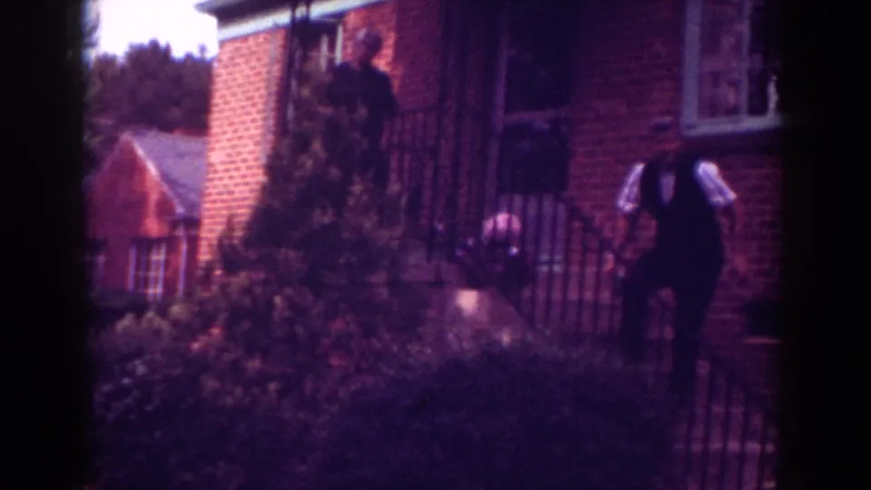 1962 Raleigh frame showing an adult in dark clothing by porch steps while another figure remains near the brick house entrance.