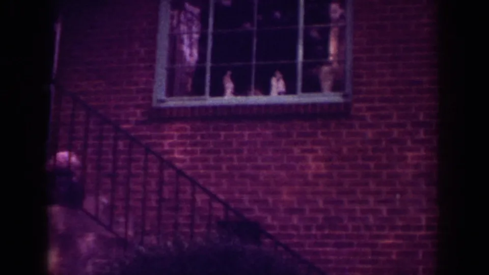The home-movie frame centers on the brick house, dark stair railing, and divided window, with faint interior shapes behind glass.