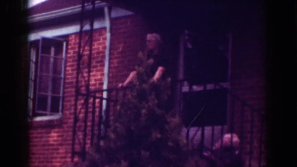 1962 Raleigh home-movie view of an elderly man on a porch, partly screened by a bush, with part of a parked car visible.