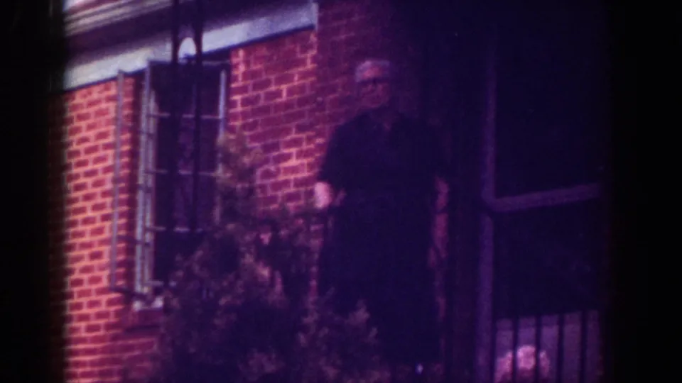 A man in a dark shirt stands by the porch of a Raleigh brick home in this 1962 home-movie frame.