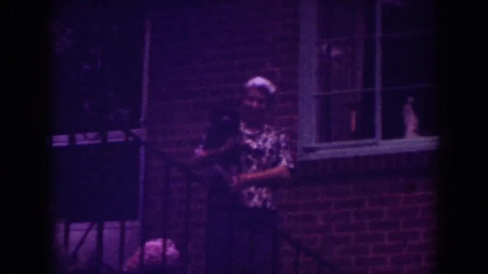 A woman stands outside the brick home while a small dog is visible near the lower porch area.
