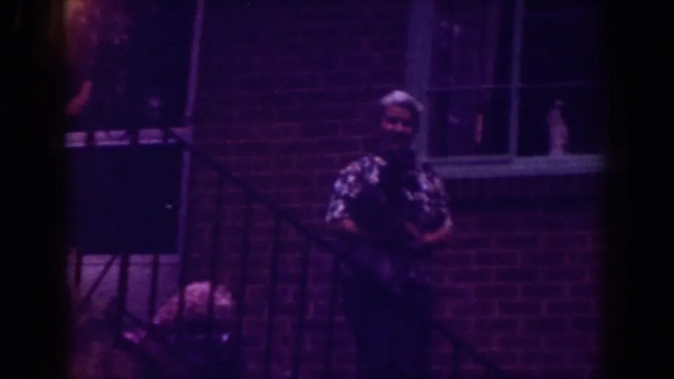The older woman faces the camera in Raleigh 1962, holding a small dark pet by an exterior stair railing against a brick building.
