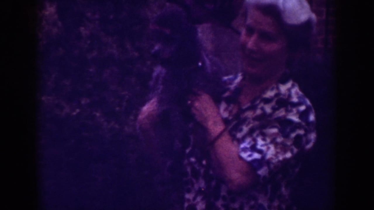 1962 Raleigh home-movie frame of a woman in a patterned blouse holding a small dark dog near greenery.
