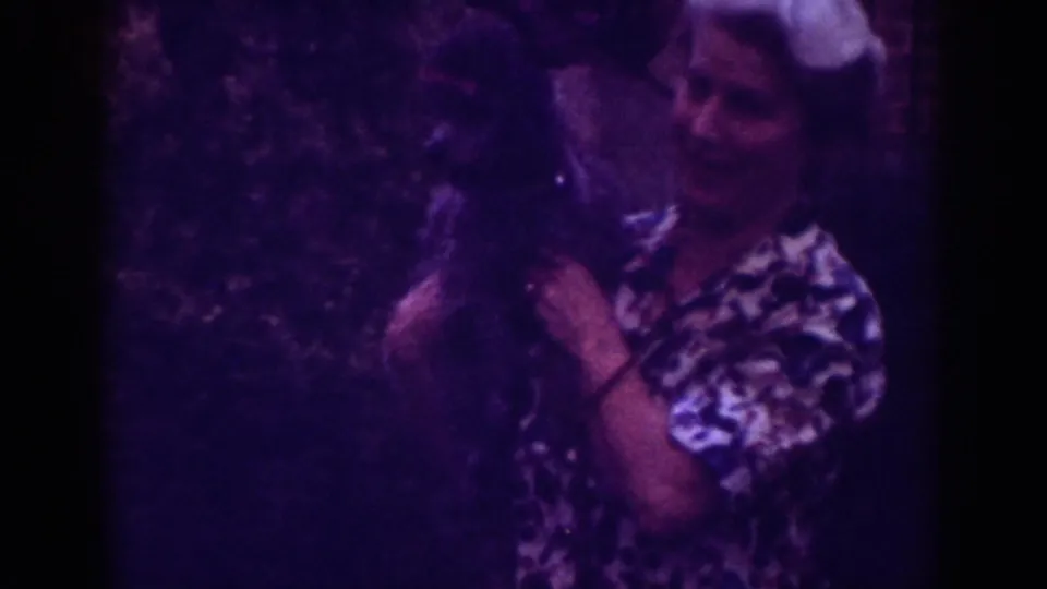 1962 Raleigh home-movie frame of a woman in a patterned blouse holding a small dark dog near greenery.