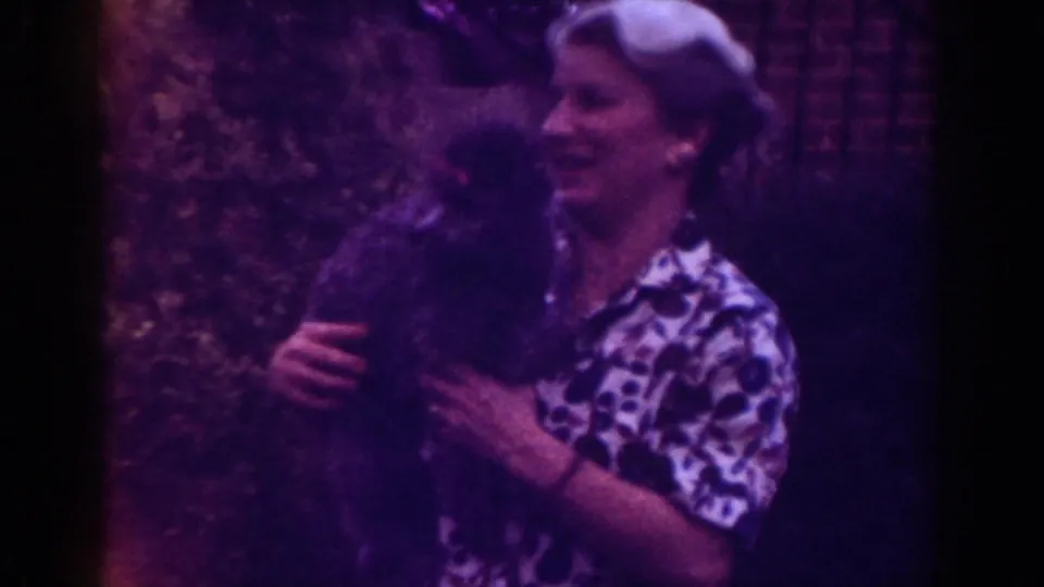 1962 Raleigh home-movie frame of a woman smiling broadly while holding a small dark dog close outdoors.