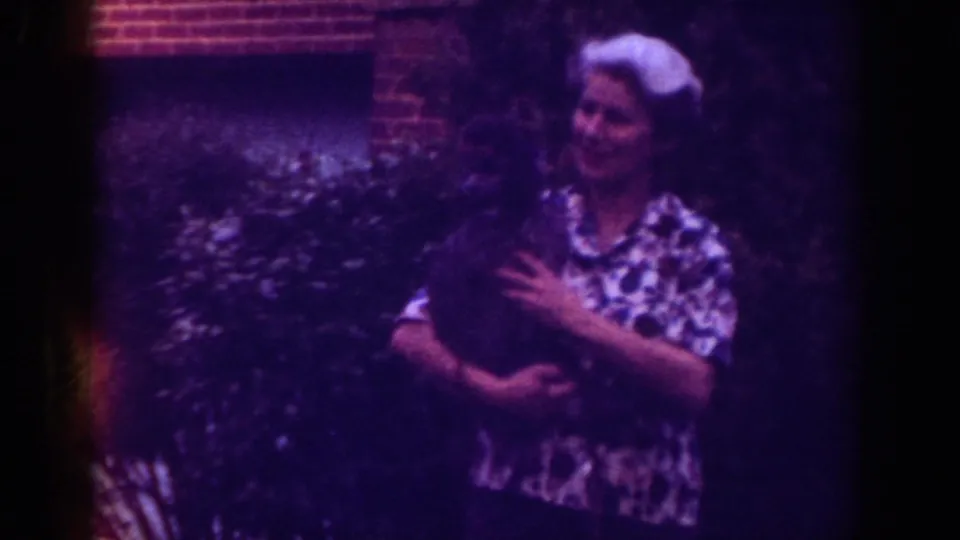 Wide 1962 Raleigh frame of a woman standing outside with a small dark dog against greenery and brick.