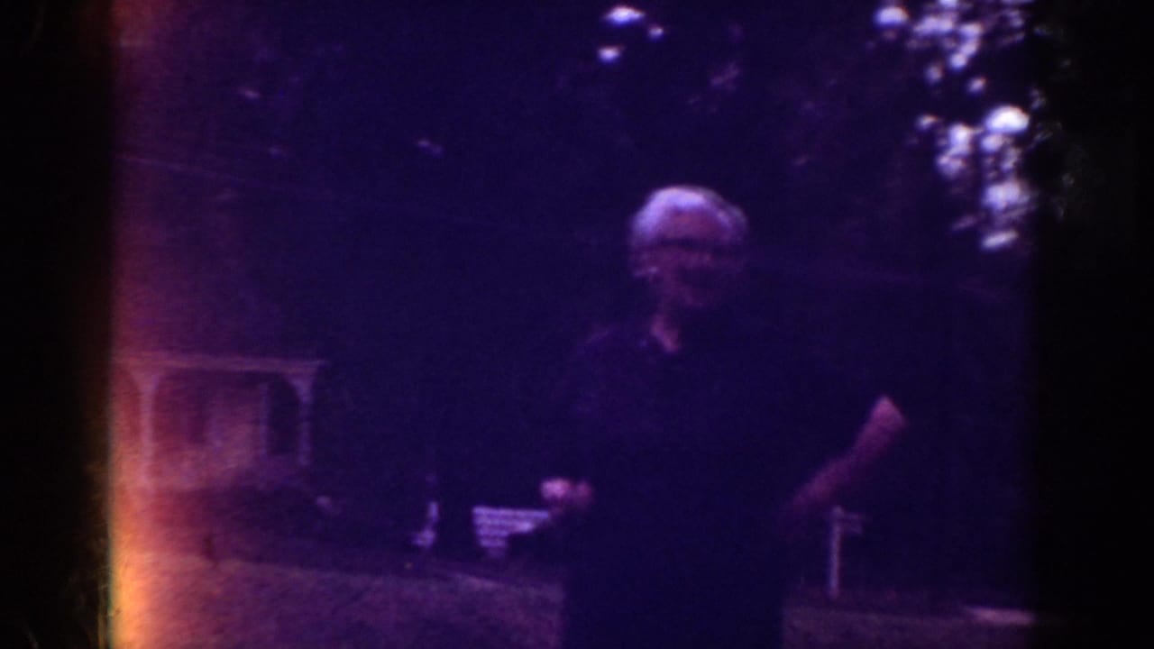 1962 Raleigh home-movie frame showing an adult in a dark shirt in a shaded residential yard with trees and lawn furniture.