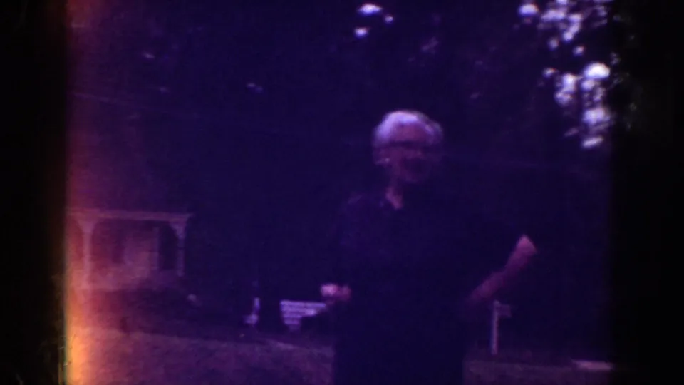 1962 Raleigh home-movie frame showing an adult in a dark shirt in a shaded residential yard with trees and lawn furniture.