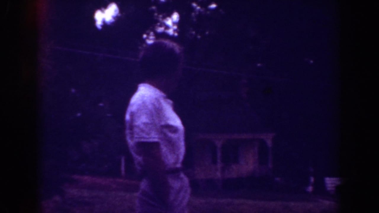 1962 Raleigh home-movie frame with an adult in a light shirt turned away in a leafy residential yard.