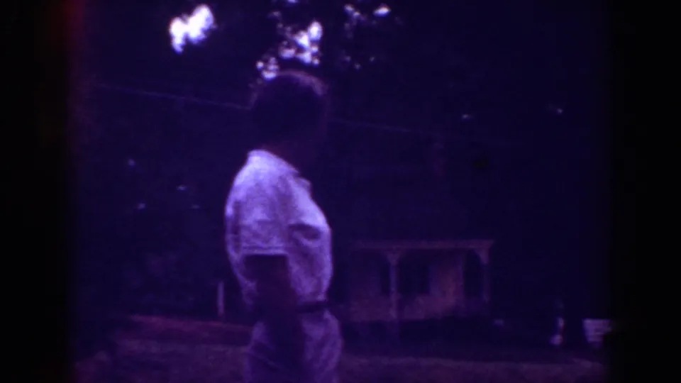 1962 Raleigh home-movie frame with an adult in a light shirt turned away in a leafy residential yard.
