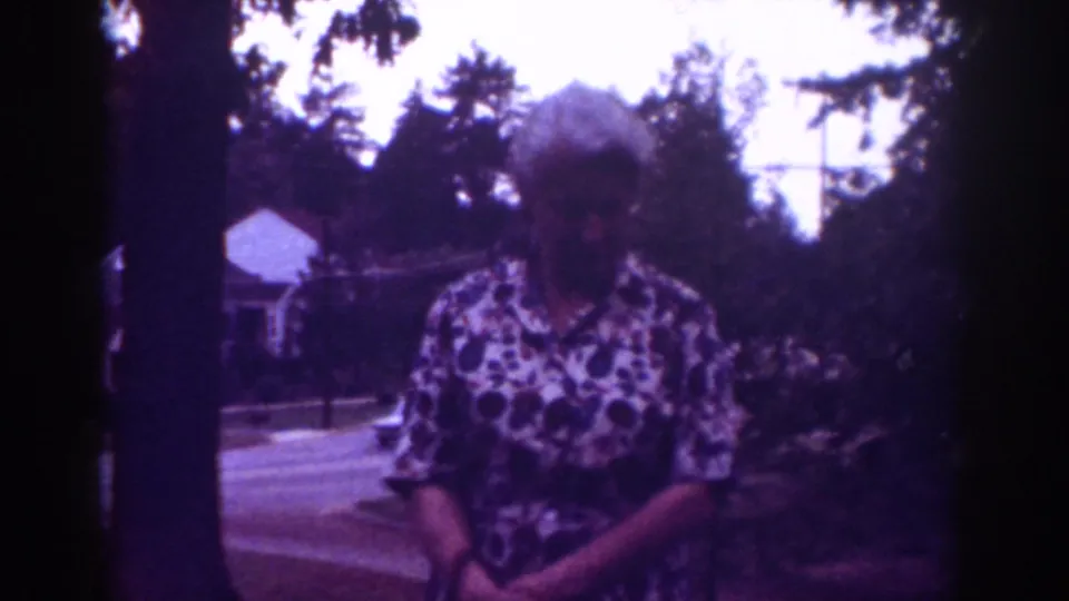 1962 Raleigh home-movie frame showing a woman in a patterned blouse holding a small dark pet near a tree-lined residential street.