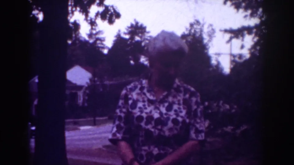 1962 Raleigh home-movie frame with a woman holding a small dark pet in a shaded yard beside trees and a residential road.