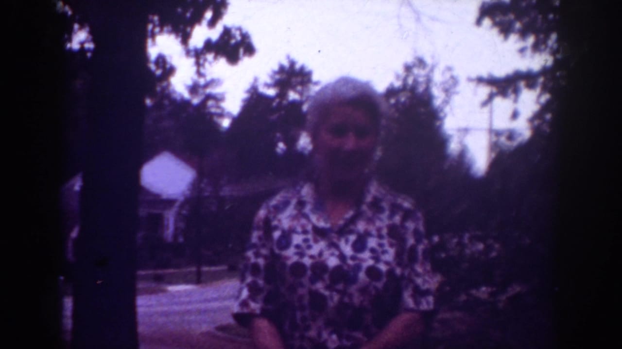 Woman in a patterned blouse stands outdoors near trees and a house.