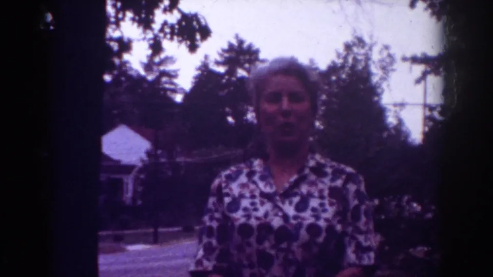 Adult woman faces the camera in a 1962 Raleigh home-movie frame, with trees and a residential background visible.