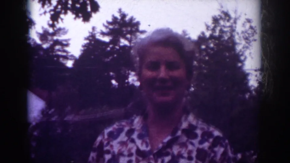 Closer 8mm view of the woman smiling outdoors in Raleigh, with trees and a residential backdrop behind her.