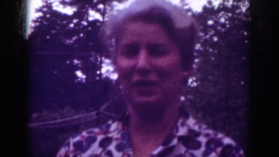 Close-up home-movie frame of the woman looking toward the camera outdoors, with dark tree shapes behind her.