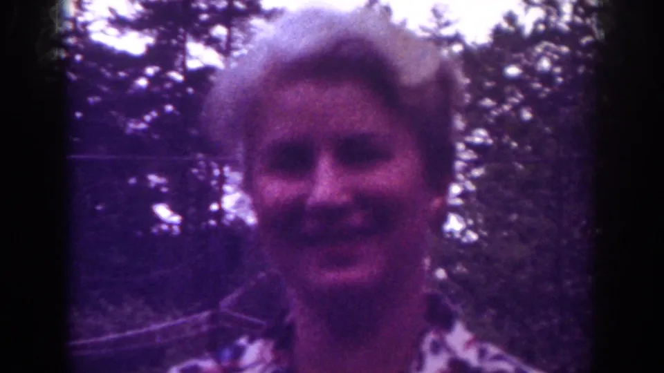In this 1962 Raleigh 8mm frame, the woman faces the camera and appears to speak, with trees and branches behind her.