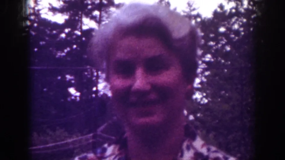 Adult blonde woman remains facing the camera in wooded 1962 Raleigh home-movie footage, with soft focus and purple film color.