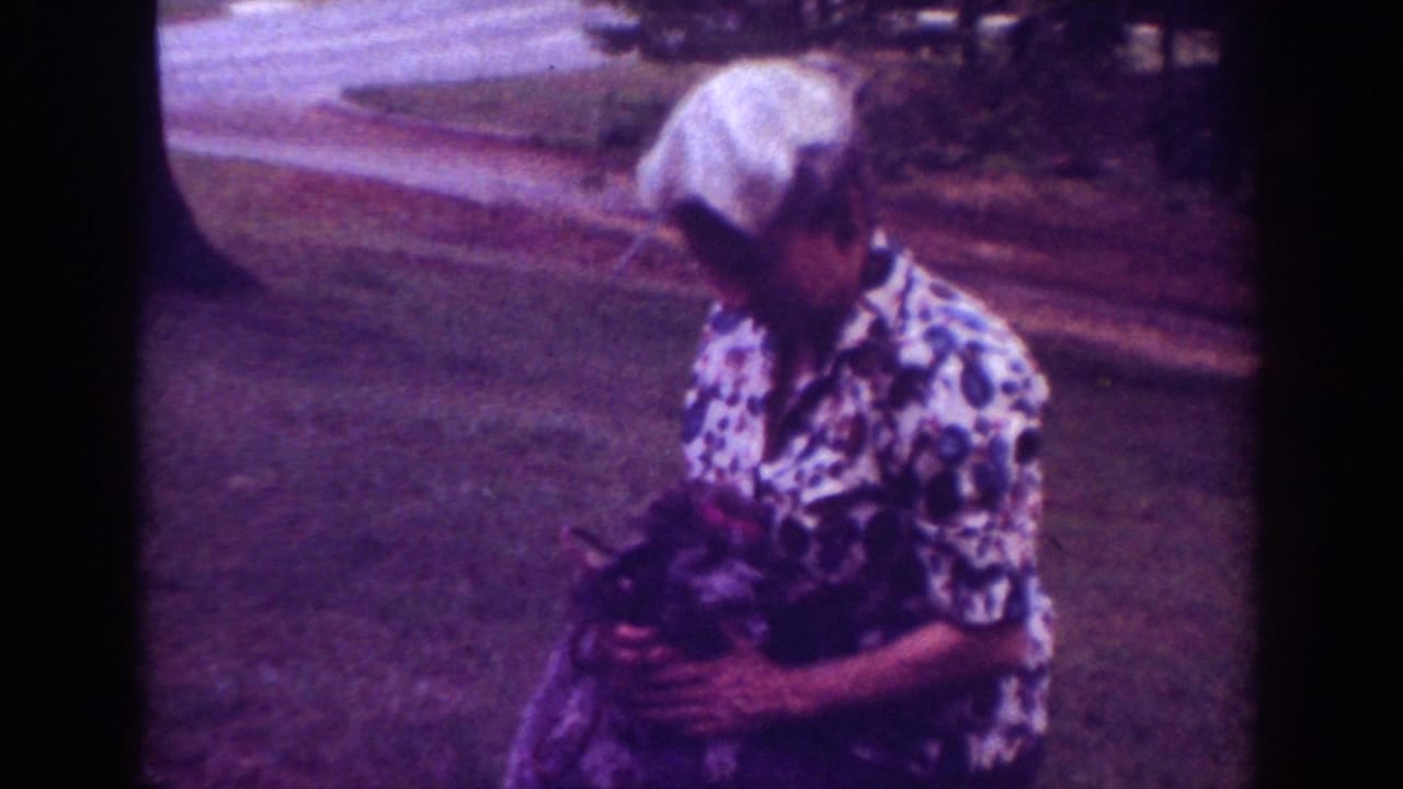 1962 Raleigh home-movie frame showing a woman with short white hair holding a small dark dog outdoors on a grassy lawn.