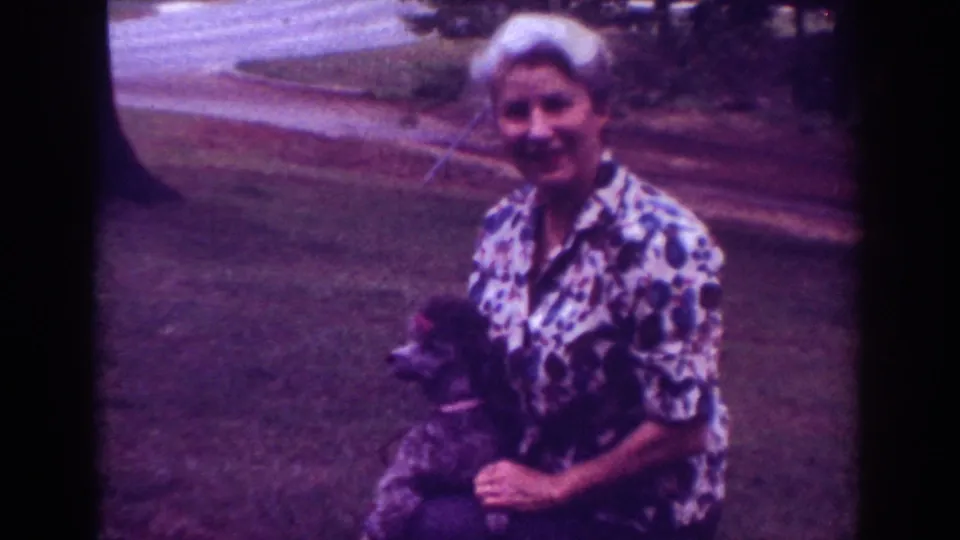 1962 Raleigh home-movie frame with the woman smiling at the camera as she holds her small dark dog on a lawn.