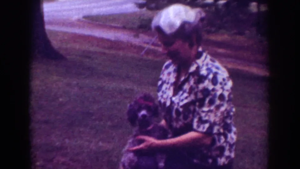 1962 Raleigh home-movie frame showing the woman looking down at her small dog while seated or crouched on the grass.