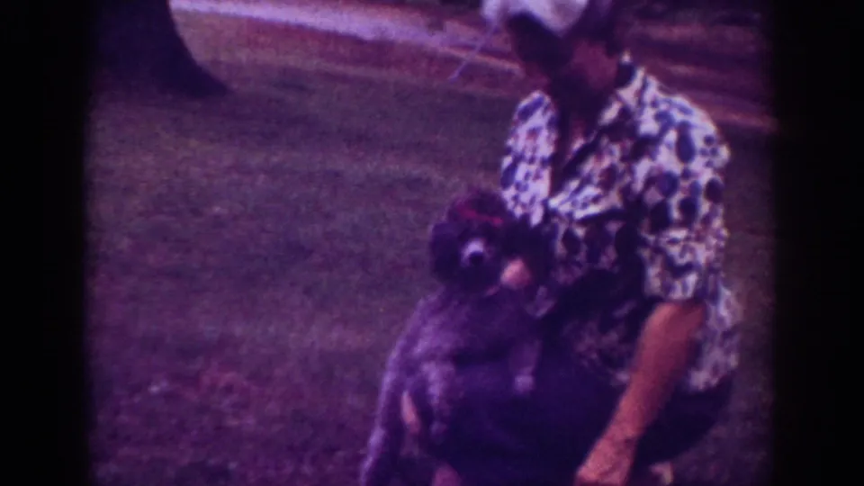 1962 Raleigh home-movie frame of the woman holding the small dark dog upright in a suburban yard with grass and trees behind her.