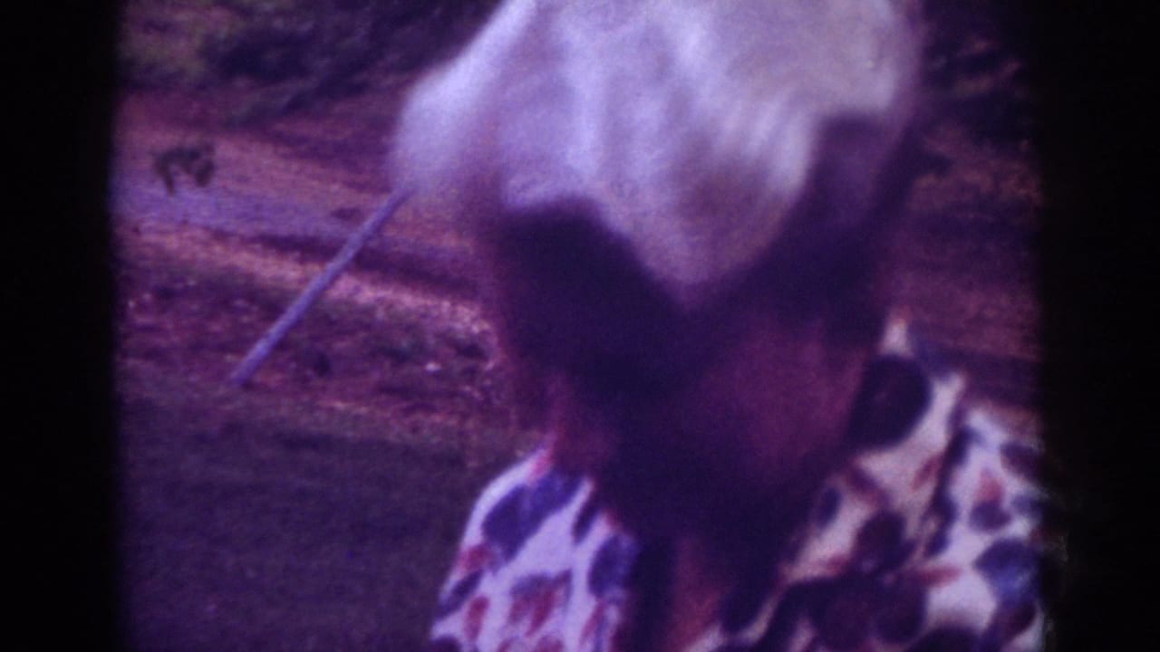 In the 1962 Raleigh 8mm footage, the older woman turns slightly while standing near grass and a path or curb.