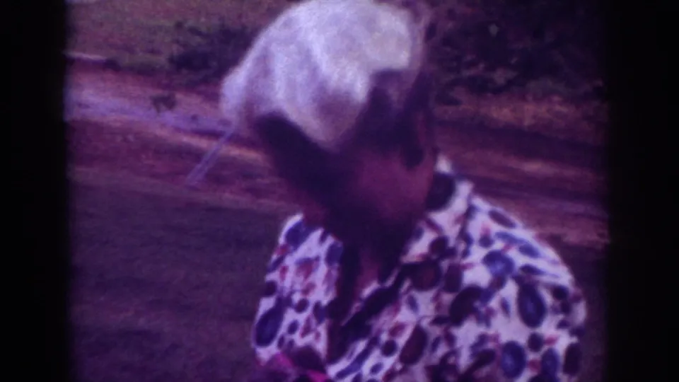 The older woman looks downward in the 1962 Raleigh home movie, with grass, a path or curb, and soft foliage behind her.