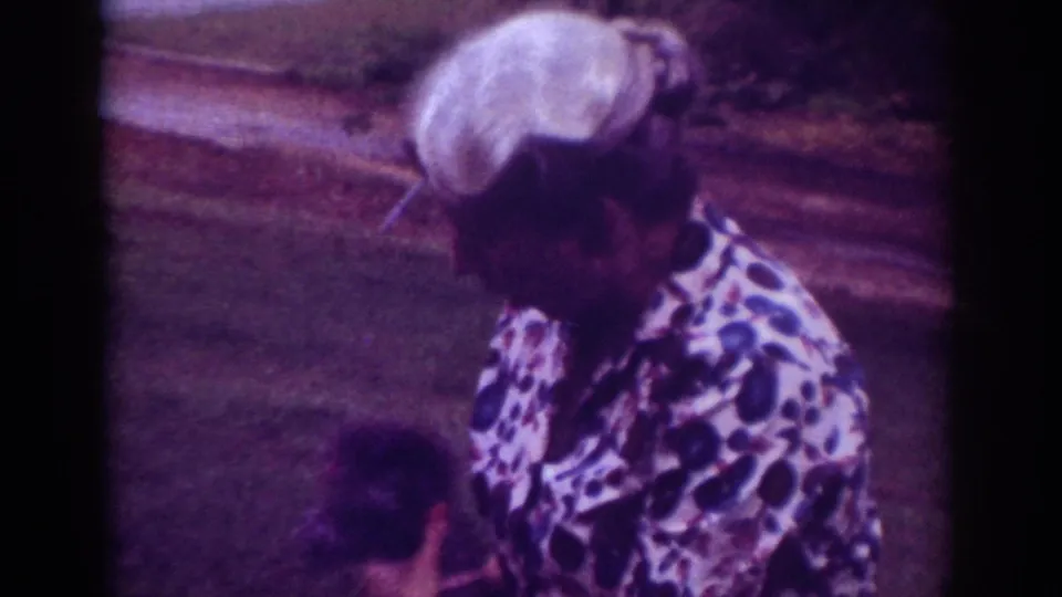 In Raleigh, 1962 home-movie footage, a woman in a patterned blouse bends toward a small dark dog near a grassy roadside.