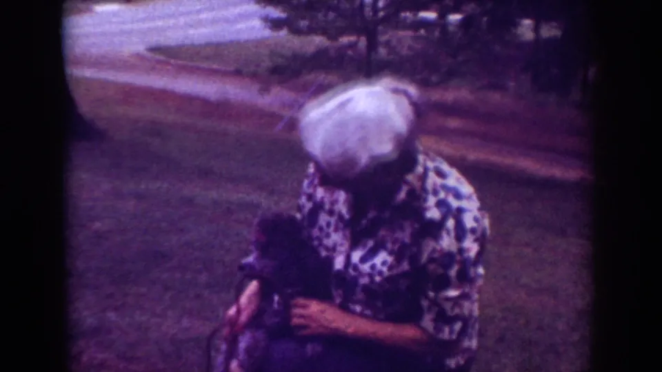 The home-movie frame shows a woman crouched on grass, interacting closely with a small dark dog near the roadside.