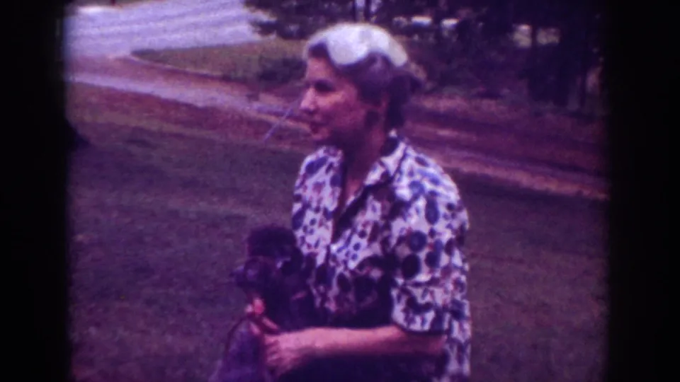 A woman in a patterned blouse holds a small dark dog on a grassy roadside in Raleigh, North Carolina, 1962.