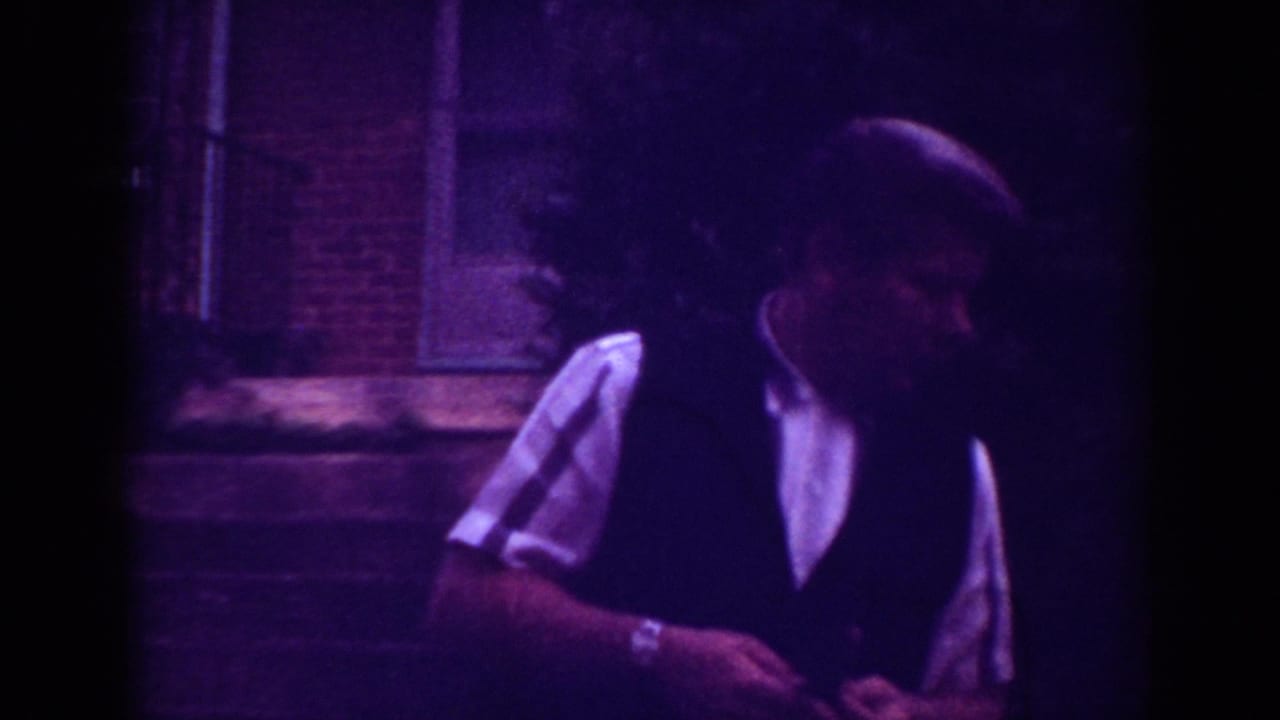 A formally dressed man stands outdoors in profile in the Raleigh home-movie scene, with brickwork and low light behind him.