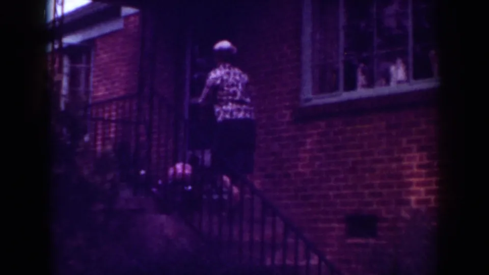 The woman continues climbing the outside staircase of a brick house in the Raleigh home-movie shot, seen from behind in soft 8mm color.